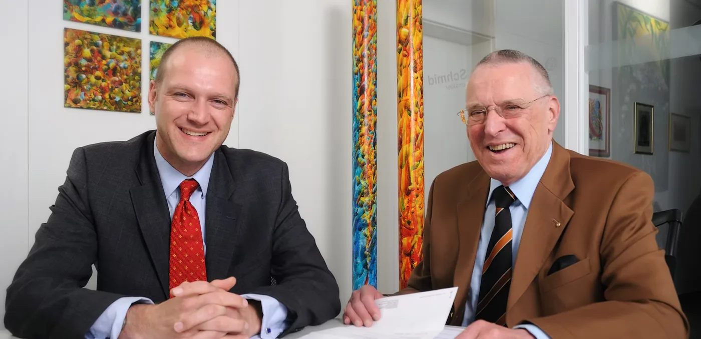 Zwei Herren im Anzug sitzen an einem Besprechungstisch, im Hintergrund Gemälde an der Wand.