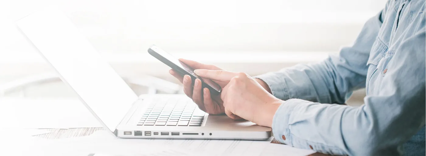 Person im hellblauen Hemd sitzt an einem Laptop und tippt auf einem Smartphone.