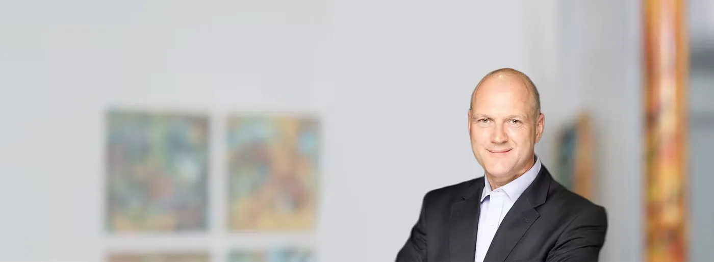 Mann im dunklen Anzug sitzt an einem Tisch, im Hintergrund eine Wand mit Gemälden.