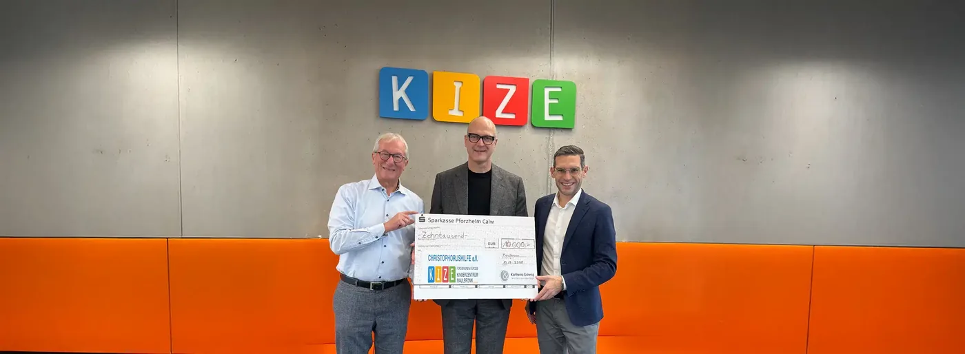 Drei Personen stehen vor einer orangefarbenen Wand mit dem Logo des Kinderzentrums KIZE und halten einen Spendenscheck.