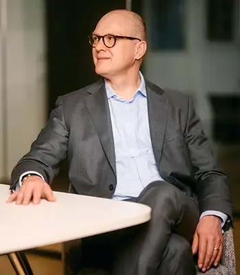 Mann mit Brille im grauen Anzug und hellblauem Hemd sitzt seitlich an einem hellen Besprechungstisch.